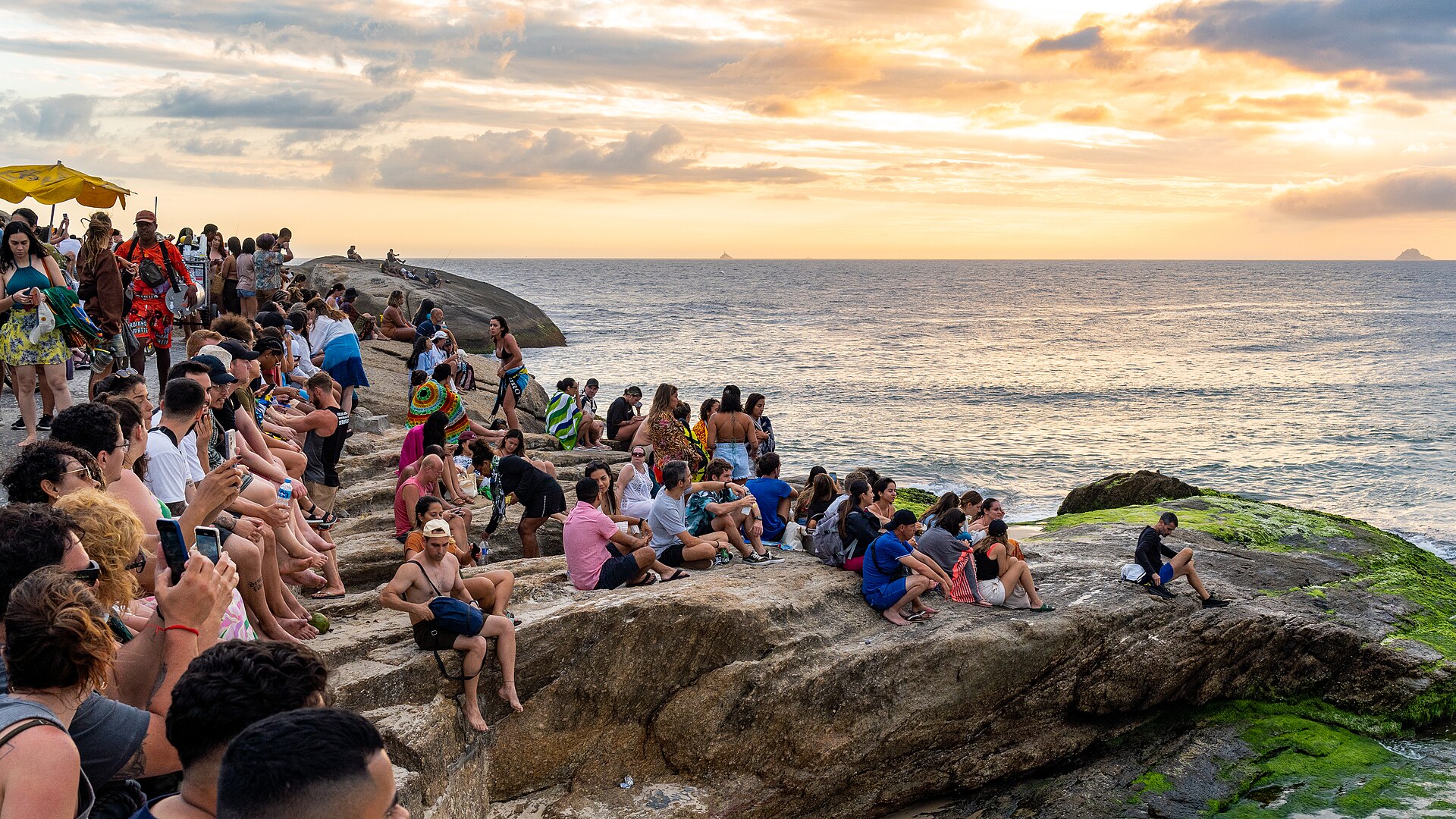 Pessoas reunidas nas pedras do Arpoador para ver o pôr do sol sobre o mar